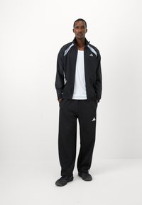 Mand står iført en sort Adidas træningsdragt med hvide striber og sorte sneakers foran en helt hvid baggrund.