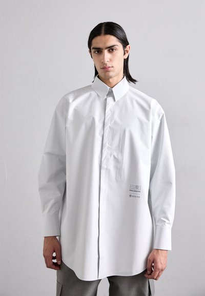 Camisa blanca de manga larga hecha de tela GORE-TEX, con cuello abotonado, bolsillo en el pecho y logotipo impreso en la parte inferior delantera.