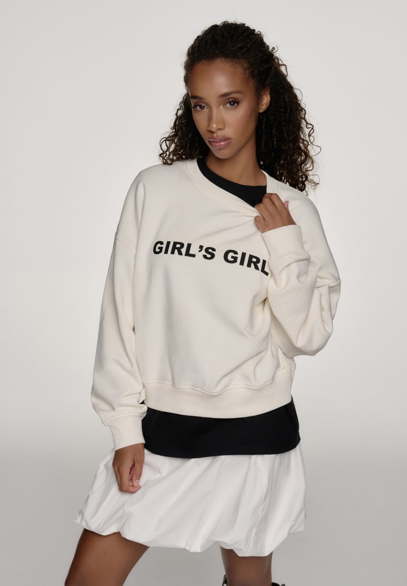 Femme aux cheveux bouclés portant un sweat-shirt blanc avec l'inscription "GIRL'S GIRL", un chemisier noir en dessous et une jupe blanche, posant devant un fond uni.