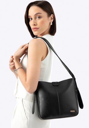 Femme aux cheveux bruns courts portant un haut blanc sans manches et tenant un sac à bandoulière en cuir noir sur fond blanc.