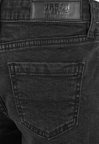 Jean en denim noir avec une étiquette en cuir portant la mention "Urban Classics". Les détails notables incluent une poche arrière avec une couture et un tissu texturé.