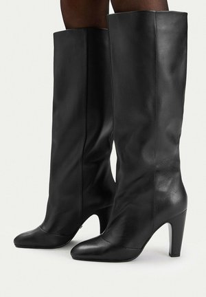 Stivali neri in pelle fino al ginocchio con punte appuntite e tacchi alti a blocco, indossati su gambe dalla pelle scura, in posizione laterale su uno sfondo semplice.