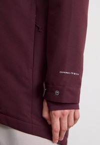 Bordeauxrote Jacke mit glatter Textur, ausgestattet mit einem verstellbaren Ärmelsaum mit Druckknopf und „OMNI-TECH“-Markierung am Ärmel.