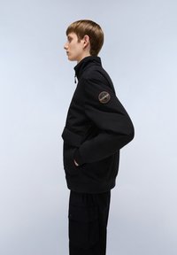 Giacca nera con zip e colletto alto, dotata di una grande tasca frontale e di una toppa rotonda sulla manica sinistra, indossata sopra pantaloni coordinati.