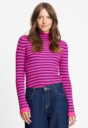 Vrouw met lang bruin haar, gekleed in een roze en paarse gestreepte coltrui en een blauwe high-waist spijkerbroek, staand tegen een witte achtergrond.