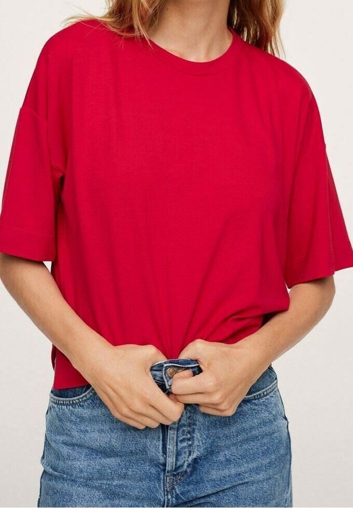 Personne portant une chemise rouge ample à manches courtes rentré dans un jean en denim bleu, en train de fermer le bouton du jean avec les deux mains.