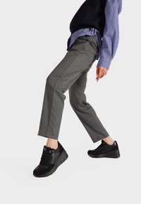 Sneakers neri con dettagli in suede texture e pelle liscia, dotati di lacci e cerniera laterale. Indossati con pantaloni grigi su misura e camicia blu.