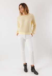 Donna con le mani in tasca, che indossa un maglione a maglia giallo chiaro, pantaloni bianchi e stivaletti neri alla caviglia, in piedi su uno sfondo bianco semplice.