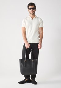Black leather tote bag with two shoulder straps and gold hardware accents, worn by a model in a light beige polo and black slim pants.