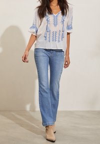 Vit broderad blus med blå mönster och tofsar, kombinerad med ljusblå utsvängda jeans och beige ankelskor.