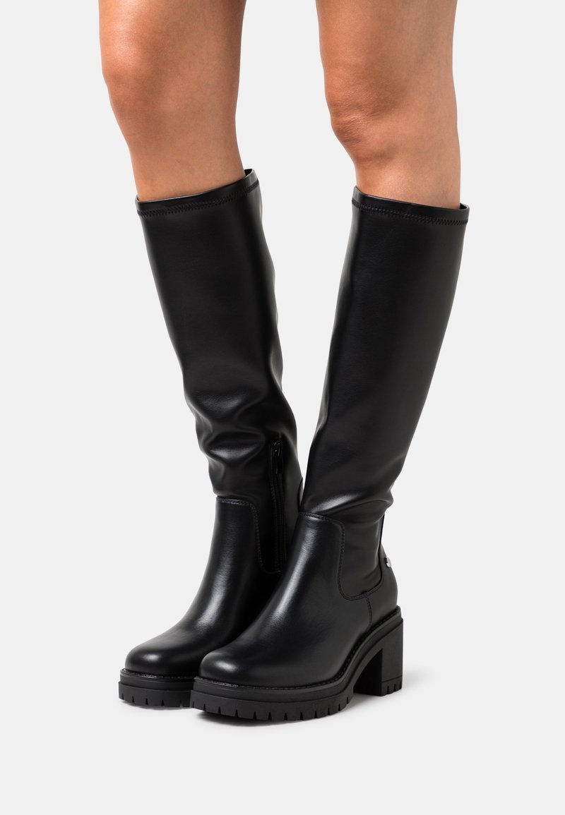 Bottes en cuir noires montant jusqu'aux genoux avec talons blocs et fermetures éclair latérales portées sur des jambes nues contre un fond uni clair.