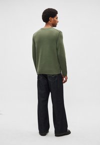 Camisola verde-oliva de mangas compridas com costuras laterais, combinada com jeans de ganga escuros de perna larga. Malha texturizada com um design simples.