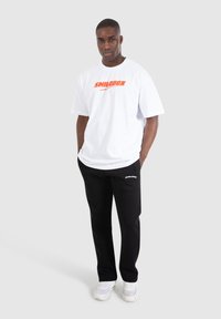 Hvid bomulds t-shirt med "SMILODON" logo i orange, kombineret med sorte sweatpants med hvidt logo på venstre lår og hvide sneakers.