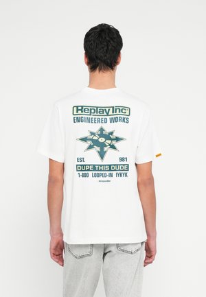 Uomo di spalle che indossa una maglietta bianca con testo grafico verde scuro "Replay Inc Engineered Works" e frecce attorno a un disco in vinile.