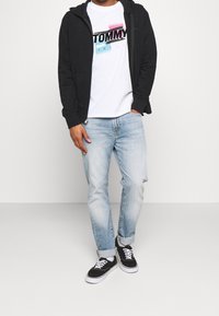 Musta vetoketjulla varustettu huppari valkoisen T-paidan päällä, jossa on värikäs "TOMMY JEANS" -logo, vaaleansiniset farkut ja mustat lenkkarit valkoisilla pohjilla.