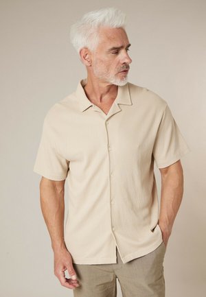 Homme aux cheveux blancs et barbe portant une chemise beige à manches courtes boutonnée et un pantalon marron clair, debout devant un fond beige uni.