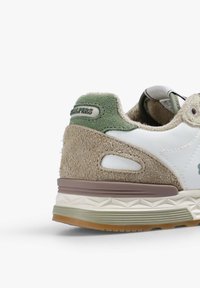Zapatilla con parte superior de cuero blanco, acentos de ante color marrón, detalles de textil verde y una suela de goma texturizada con un diseño de varias capas.
