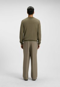 Mann mit kurzem dunklen Haar steht mit dem Rücken zu uns, trägt ein olivgrünes Langarmshirt und lockere beigen Hosen vor einem einfarbigen Hintergrund.