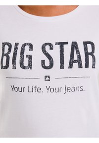 Valge T-särk tuhmunud musta tekstiga "BIG STAR" üle "Your Life. Your jeans." kandja kaela ja õlgadega.