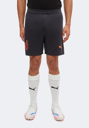 Homme portant un short de sport noir Puma avec le logo de l'AC Milan, des chaussettes blanches Puma et des chaussures de football blanches avec des touches bleues et rouges.