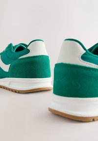 Baskets en daim vert et textile avec des accents blancs, dotées d'une semelle en caoutchouc et d'un col rembourré, vues sous un angle arrière.