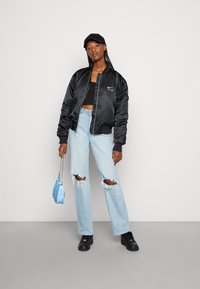 Giacca bomber nera in satin con polsini a costine, abbinata a jeans strappati blu chiaro. In mano tiene una borsa a tracolla blu con motivo, completando il look con sneakers nere.
