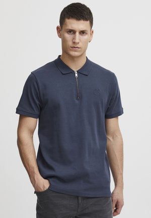 Marineblå poloshirt med glidelås i kragen, laget av glatt stoff. Har korte ermer og en subtil logo på brystet. Tilpasset passform.
