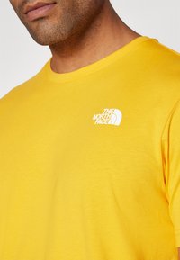 Gelbes T-Shirt mit kurzen Ärmeln aus Baumwolle, mit Rundhalsausschnitt, mit einem weißen Logo von "The North Face" im linken Brustbereich.
