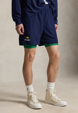 Hombre vistiendo pantalones cortos azul marino US Open sobre pantalones cortos de compresión verdes, calcetines blancos y zapatillas altas color blanco hueso.