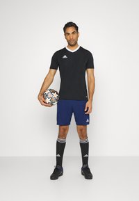 Camiseta deportiva negra con ribetes blancos, pantalones cortos azules, calcetas de fútbol negras con rayas blancas y zapatos de fútbol negros, sosteniendo una pelota con diseño.