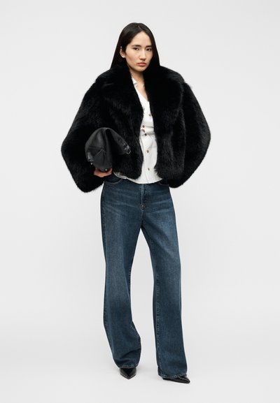 Μαύρο μπουφάν από συνθετική γούνα, λευκό πουκάμισο με κουμπιά, μπλε τζιν φαρδιάς γραμμής, μαύρο τσαντάκι χειρός και μαύρες μυτερές γόβες, πάνω σε ουδέτερο φόντο.