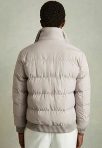 Jachetă puffer de culoare gri deschis, cu guler înalt, tiv și manșete ribbate, având secțiuni orizontale matlasate pentru izolație.