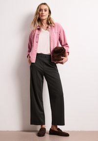 Femme debout contre un mur, portant une veste rose, un haut blanc, un pantalon large noir, des mocassins marron et tenant une pochette marron.