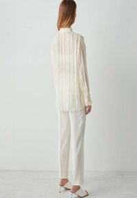 Blouse crème clair plissée avec col montant, manches longues et poignets à boutons, associée à un pantalon blanc ajusté et des chaussures à talons ouverts.