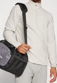 Muž oblečený do světle béžového svetru Champion na čtvrtinový zip a šedých tepláků, držící černou a šedou tašku přes rameno s síťovanou kapsou.