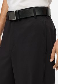 Vīrietis, kurš valkā melnus bikses ar melnu ādas jostu un baltu kreklu, roka atpūšas blakus kabatai uz baltā fona.