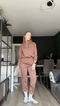 Smeđi trenirka s gornjim dijelom na pola patentnog zatvarača i hlačama s elastičnom gumom. Bijeli sportski tenisice s crnim prugama i bijelim čarapa do gležnja. Unutarnje okruženje.