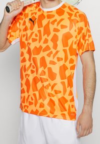 Camiseta de manga corta naranja con un patrón de jirafa, que presenta un logo negro y detalles blancos en las mangas. Material transpirable.
