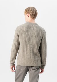 Pull en tricot beige avec des manches longues, encolure ronde et poignets côtelés. Porté avec un pantalon gris, mettant en avant une texture intéressante.