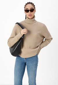Suéter de punto beige con cuello alto, combinado con vaqueros azules y un bolso de hombro de cuero negro. Los accesorios incluyen gafas de sol de ojo de gato y pendientes de aro dorados.