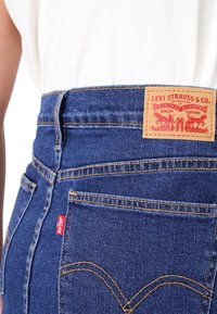 Blue denim jeans with a high waist, featuring a leather Levi Strauss tag, contrasting yellow stitching, and a small red logo on the back pocket.