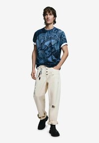 Modrá vzorovaná košeľa s krátkym rukávom, krémové džínsy s opotrebovaným vzhľadom a zahnutými manžetami, a čierne členkové čižmy. Detaily zahŕňajú gombíky a zipsy.