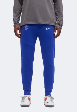 Άτομο που φοράει μπλε φόρμα Nike Paris Saint-Germain και λευκά αθλητικά παπούτσια Nike με κόκκινα σχέδια καρδιών, στέκεται με χαλαρά χέρια.
