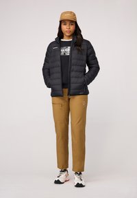 Svart pufferjacka med dragkedja fram, bar över en svart sweatshirt med grafiskt tryck; beige byxor och beige keps; sportiga vita skor.
