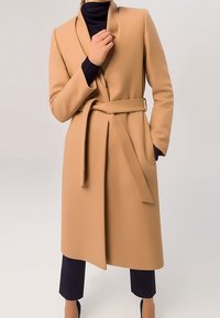 Abrigo beige texturizado con un collar ancho, cinturón en la cintura y mangas largas, combinado con una camiseta oscura y pantalones cortos.
