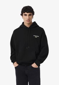 Joven con cabello rizado oscuro que lleva una sudadera negra con "Jeu Set et Match" y un logo de cocodrilo verde, con las manos en los bolsillos, fondo neutro.