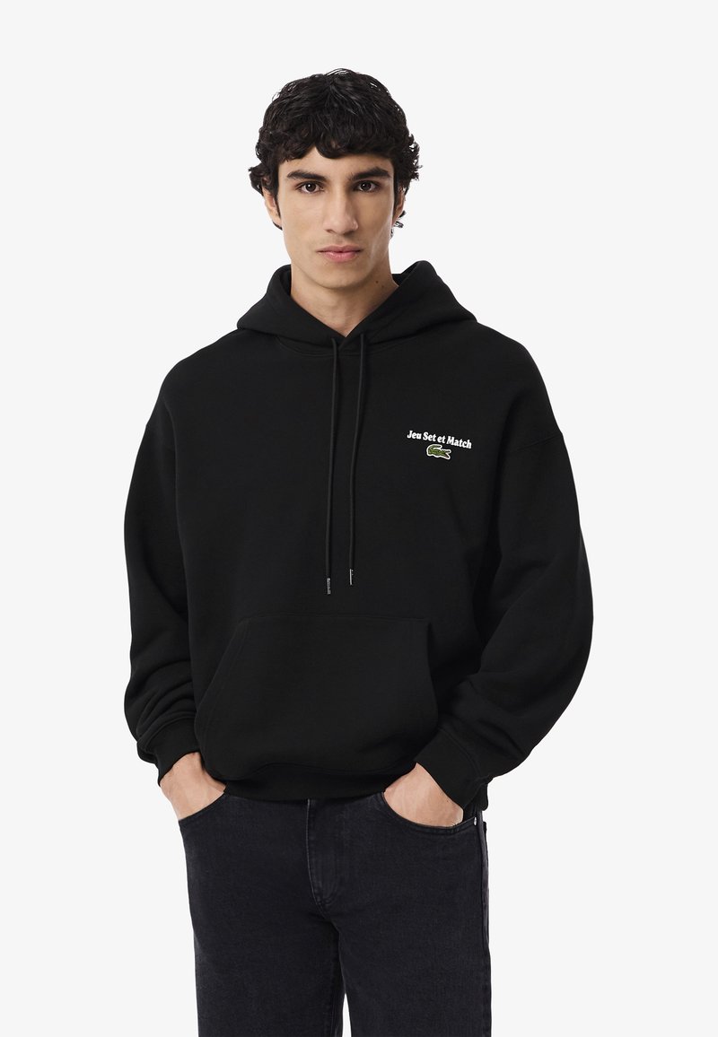 Joven con cabello rizado oscuro que lleva una sudadera negra con "Jeu Set et Match" y un logo de cocodrilo verde, con las manos en los bolsillos, fondo neutro.
