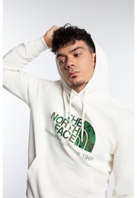 Sudadera color crema hecha de material suave que presenta un gran logotipo gráfico verde con textura de camuflaje y un bolsillo tipo canguro. Incluye cordones.