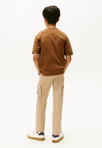 Korte bruine shirt met korte mouwen en een kleine vlagaccent, gecombineerd met beige cargobroeken met zijzakken en witte sneakers met tan kleurige zolen.