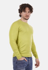 Uomo con capelli ricci che indossa un maglione giallo a maniche lunghe e jeans blu, in piedi con una mano in tasca su uno sfondo bianco.